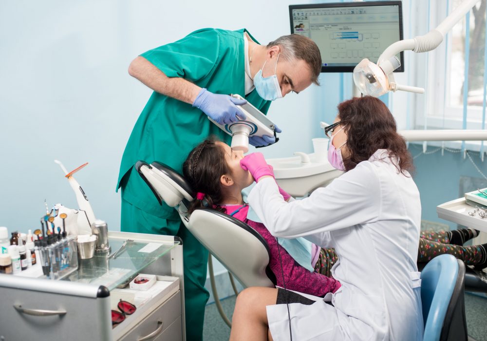 Senior pediatric dentist with nurse doing dental treatment patient girl using dental x-ray machine in dental office. On the background monitor with X-ray the patient's teeth. Dental equipment